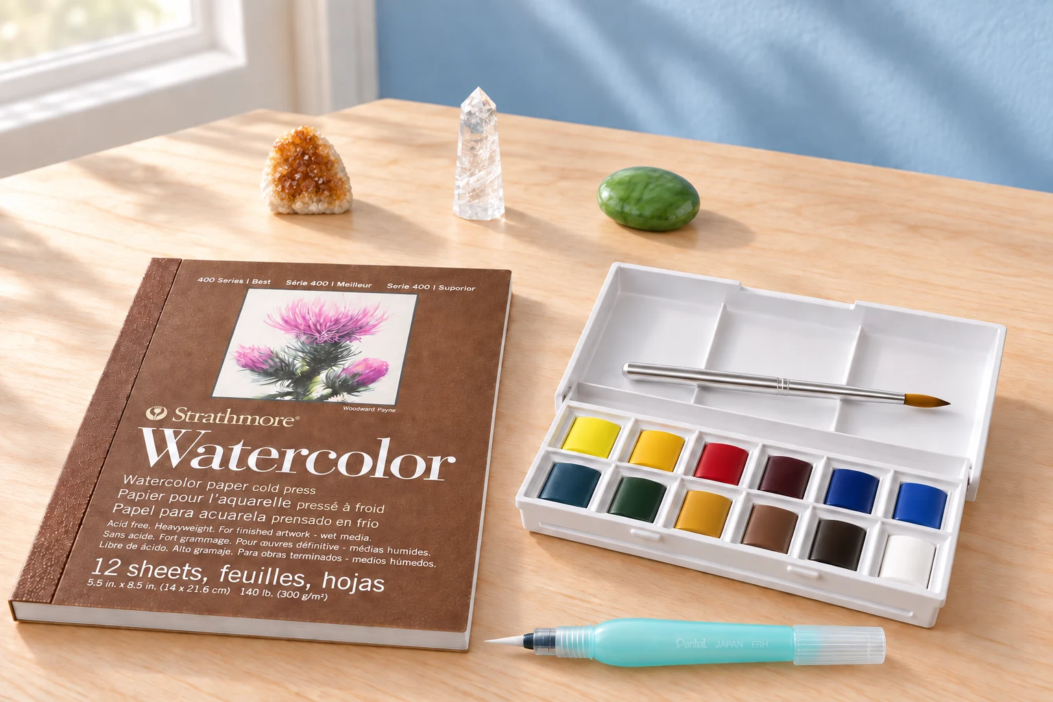 A watercolor pad, open twelve-pan pocket palette and aqua water brush with citrine, aventurine and clear quartz on a sunlit craft table.