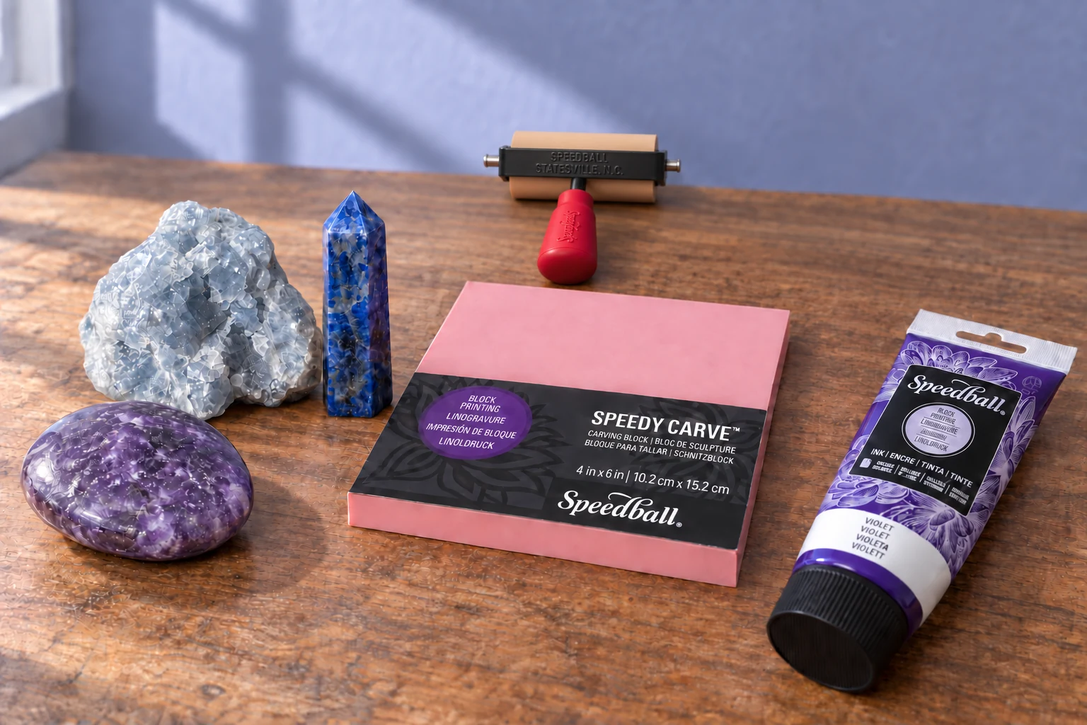 A lilac lepidolite palm, rough blue calcite and sodalite point beside a pink block, red handled brayer and capped violet printing ink.