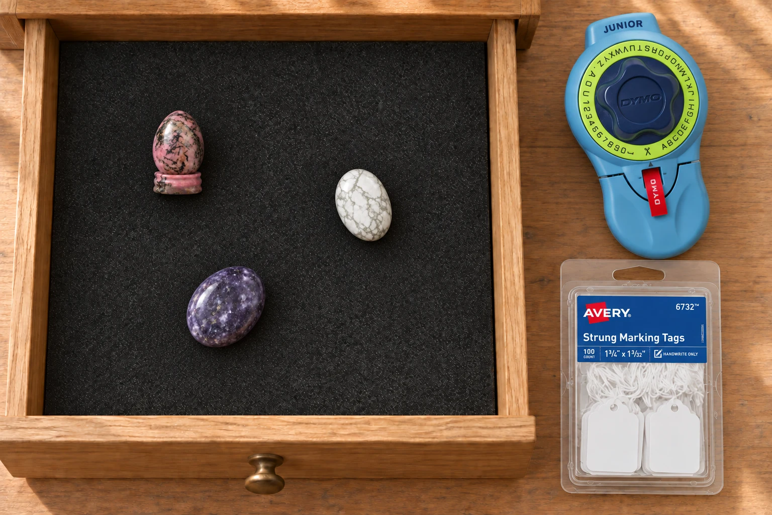 Pink-black rhodonite egg, white howlite and purple lepidolite on black foam in an open oak drawer, with a blue DYMO labeler and Avery strung-tag package beside it.