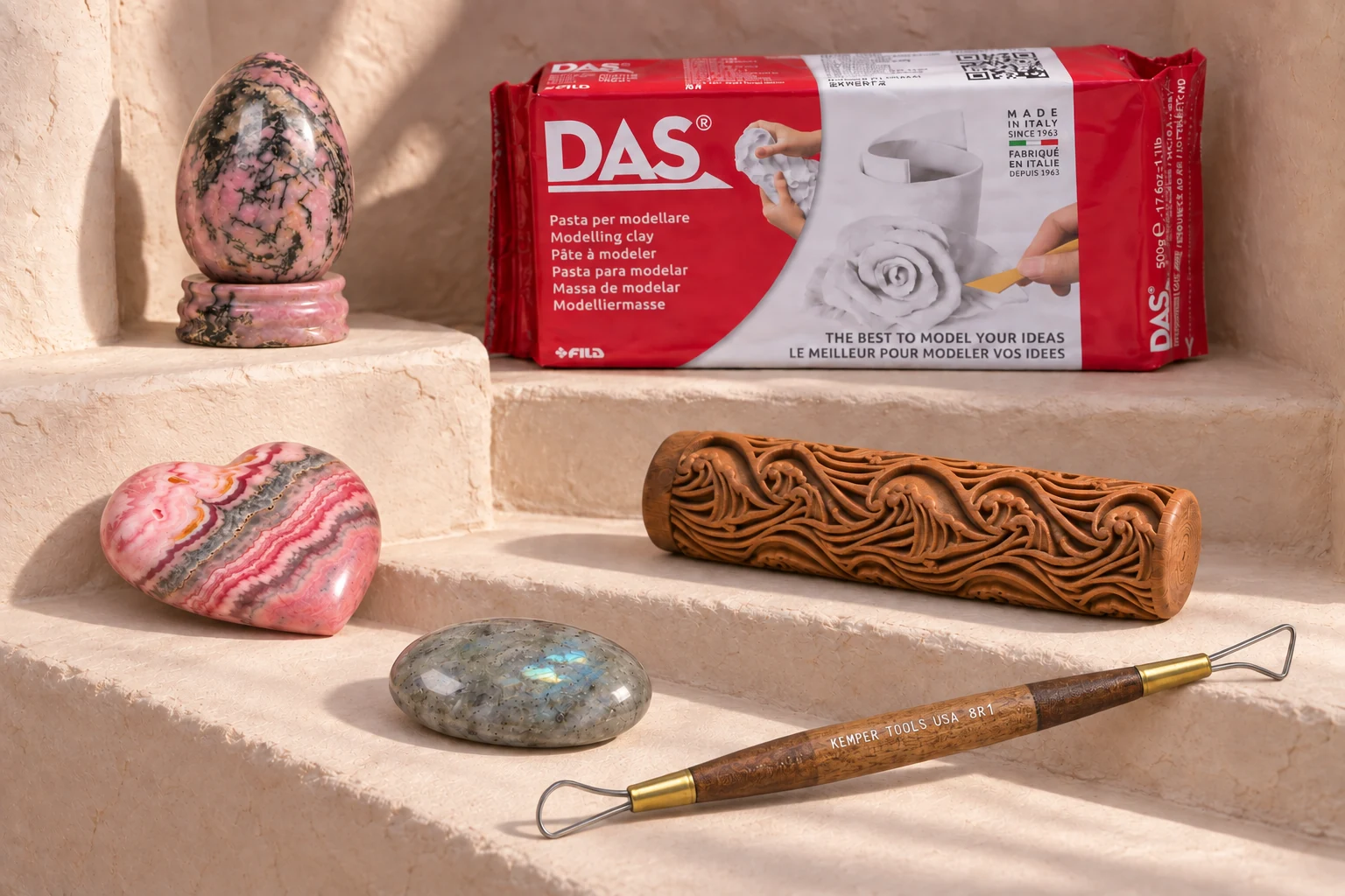 In a warm plaster alcove, a rhodonite egg on its stand, banded rhodochrosite heart and gray moonstone sit at staggered levels with wrapped DAS clay, a carved wooden Waves roller and a double-loop ribbon tool.