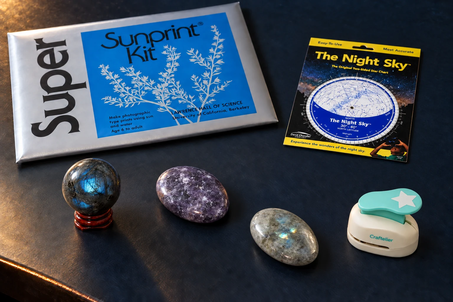 On a midnight desk, a blue-flashed labradorite sphere on its wood stand, lilac lepidolite and gray moonstone sit below a sealed Sunprint Kit and rotating Night Sky chart, with a turquoise star punch to one side.